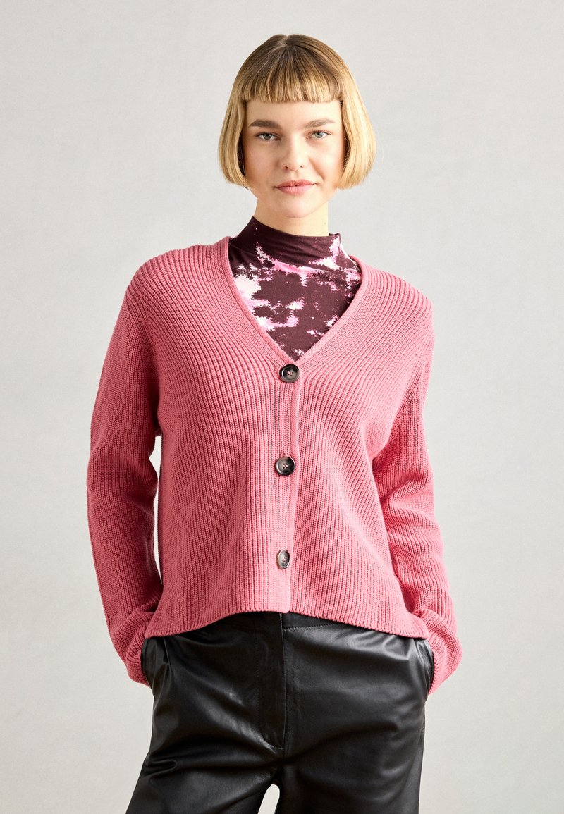 Rožinė megzta liemenė su V formos iškirpte ir penkiais tamsiais mygtukais, turinti raštuotą tekstūrą ir ilgomis rankovėmis, uždėta ant rudos blykstės marškinėlių.