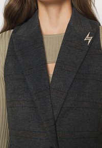Gilet grigio in lana con collarino a scollo a V, sottile motivo a quadri e una spilla decorativa a forma di fulmine. Tessuto texturizzato con una vestibilità su misura.