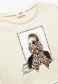 Creme sweatshirt med rund halsudskæring, der har et trykt billede af en piges ryg med en leopardmønstret hårsløjfe og tekst nedenfor.