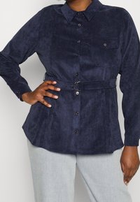 Marinblå skjorta med knappar i mjuk mockaliknande tyg, med långa ärmar, krage, framficka och midja med bälte och metallring som accent.