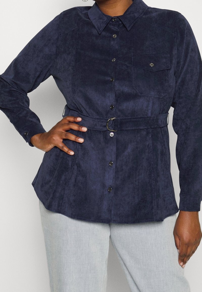 Marinblå skjorta med knappar i mjuk mockaliknande tyg, med långa ärmar, krage, framficka och midja med bälte och metallring som accent.