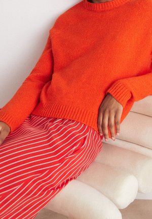 Personne portant un pull en tricot orange vif et une jupe rouge à rayures verticales blanches, assise sur une chaise rembourrée beige clair.