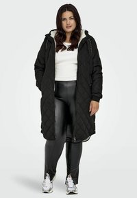 Schwarzer, gesteppter langer Mantel mit Kapuze und weißem Fleece-Futter, getragen über einem weißen Oberteil und glänzenden schwarzen Leggings. Verfügt über einen Frontreißverschluss und Druckknöpfe.