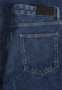 Vaqueros de mezclilla en azul oscuro con un acabado texturizado. Incluye costuras amarillas en contraste y un parche de cuero negro en la cintura.