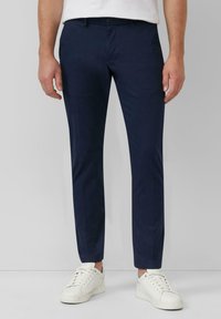 Muž v úzkych tmavomodrých nohaviciach a bielych teniskách, stojí na jednoduchom svetlosivom pozadí.