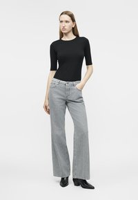 Žena oblečená v čiernom top s dĺžkou rukáva po lakeť, svetlošedé široké rifle s rukou v kapse a čierne členkové topánky na opätku, stojaca na bielom pozadí.