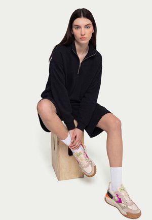 Jeune femme assise sur une caisse en bois, portant une robe noire à manches longues avec fermeture éclair, des chaussettes blanches et des baskets beiges avec des accents colorés.