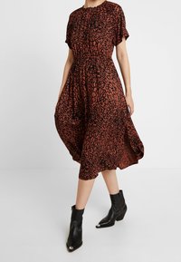 Langes, locker sitzendes Kleid in Rost mit schwarzem Leopardenmuster. Verfügt über einen gerafften Halsausschnitt und kurze Ärmel. Kombiniert mit schwarzen Knöchelstiefeln.