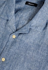 Camicia in lino azzurro chiaro con colletto a polo e due bottoni beige. Il tessuto presenta una leggera trama testurizzata e un'aspetto leggermente stropicciato.