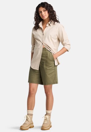 Chemise beige à boutons, shorts verts et bottes en cuir beiges à lacets avec des semelles texturées. Les chaussettes ont des rayures légères. Le modèle se tient avec les mains dans les poches.
