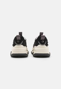 Baskets noires et beiges avec semelle épaisse, tige en mesh et en daim, et languettes de tirage à l'arrière, présentées de dos sur fond blanc.