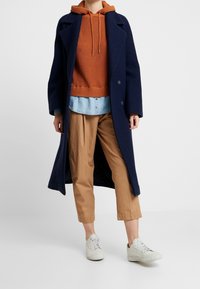 Laivastonsininen pitkä takki, jossa on lovettu kaulus, yhdistettynä oranssiin ribbineulottuun huppariin, vaaleaan farkkupaitaan ja beigen värisiin lyhytlahkeisiin housuihin. Valkoiset tennarit täydentävät asun.