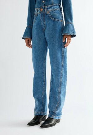 Persoon draagt mediumblauwe high-waist rechte jeans met een bijpassend spijkerjasje en zwarte enkellaarsjes met een puntige neus.