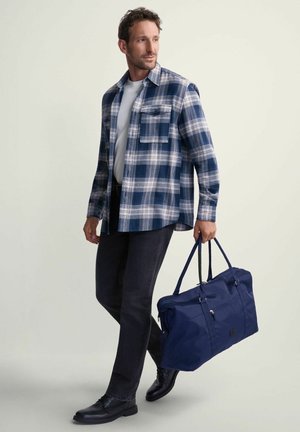 Koszula flanelowa w niebiesko-białą kratę, ciemne dżinsy, skórzane buty oraz granatowa torba typu duffel z podwójnymi uchwytami, noszone przez mężczyznę.