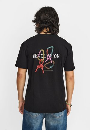 Persona con capelli neri fino alle spalle indossa una maglietta nera con moschettoni da arrampicata colorati e la scritta "REVOLUTION" sulla schiena.