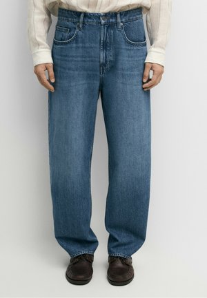 Mand iført løstsiddende blå jeans, hvid langærmet skjorte og mørkebrune sko mod en ensfarvet baggrund.