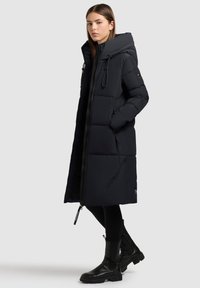 Lang sort pufferjakke med høj krave, frontlynlås og snoretræk i hætten. Har et quiltet design og sidelommer.