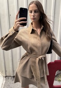 Vestido camisa de ante en tono marrón claro con cintura ceñida y cinturón, que presenta un cuello camisero, detalles de botones y una tela texturizada. Los accesorios incluyen un bolso de punto.