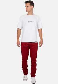 T-shirt bianca in cotone con maniche raglan corte e logo nero. Abbinata a pantaloni rossi in cotone e sneaker bianche. Design semplice e casual.