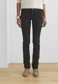 Jeans negros ajustados con una textura suave, con bolsillos frontales y combinados con mocasines color marrón claro. El modelo lleva un suéter de color claro.