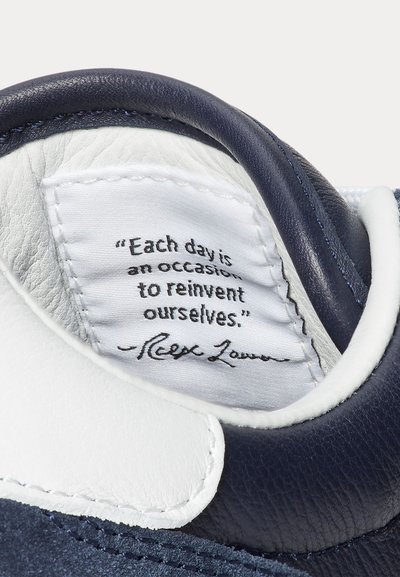 Tengerészkék bőrcipő belseje, fehér bőrkiegészítővel és egy címkével, amely fekete betűkkel motiváló idézetet tartalmaz. Sima textúra.