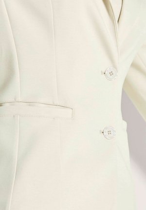 Cremfarbener Blazer mit zwei runden, transparenten Knöpfen und einer horizontalen Schlitztasche auf der linken Seite, die detaillierte Nähte zeigt.