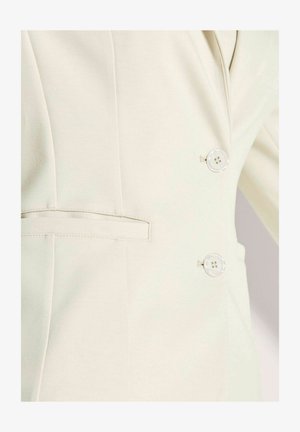 Cream-colored blazer with two round translucent buttons and a horizontal slit pocket on the left side, showing detailed stitching.