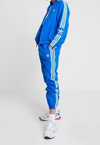 Chándal azul con franjas blancas, que incluye una chaqueta con cremallera y pantalones con cinturilla elástica. Llevado con zapatillas blancas con acentos coloridos.