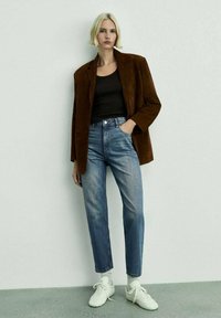 Rjav suedni blejzer, črn rebrast top z naramnicami, svetlo modri hlače z visokim pasom, bele superge, stoječa na sivi tleh pred enostavno ozadje.