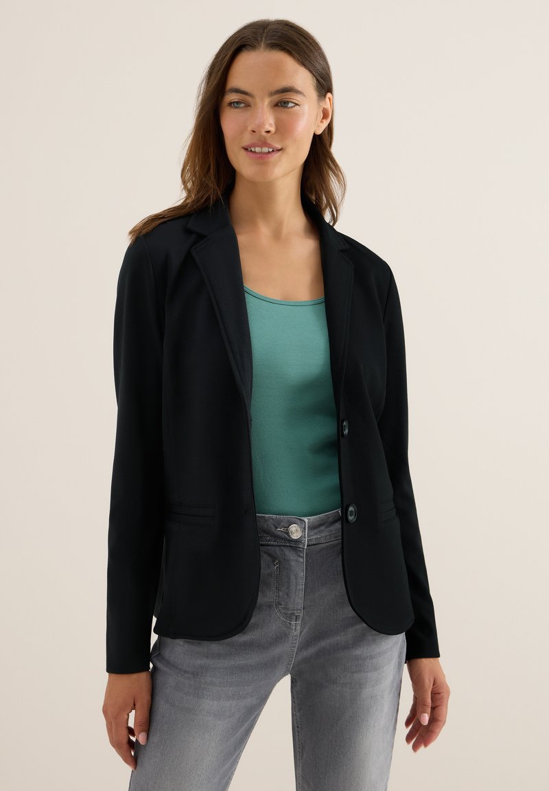 Vrouw in een zwarte blazer over een teal tanktop en grijze jeans, staand met ontspannen armen tegen een effen beige achtergrond.