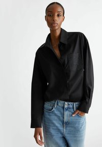 Camisa negra de manga larga con cierre de botones, que presenta un bolsillo texturizado con tachuelas. Se lleva con jeans de tiro alto de color azul claro.