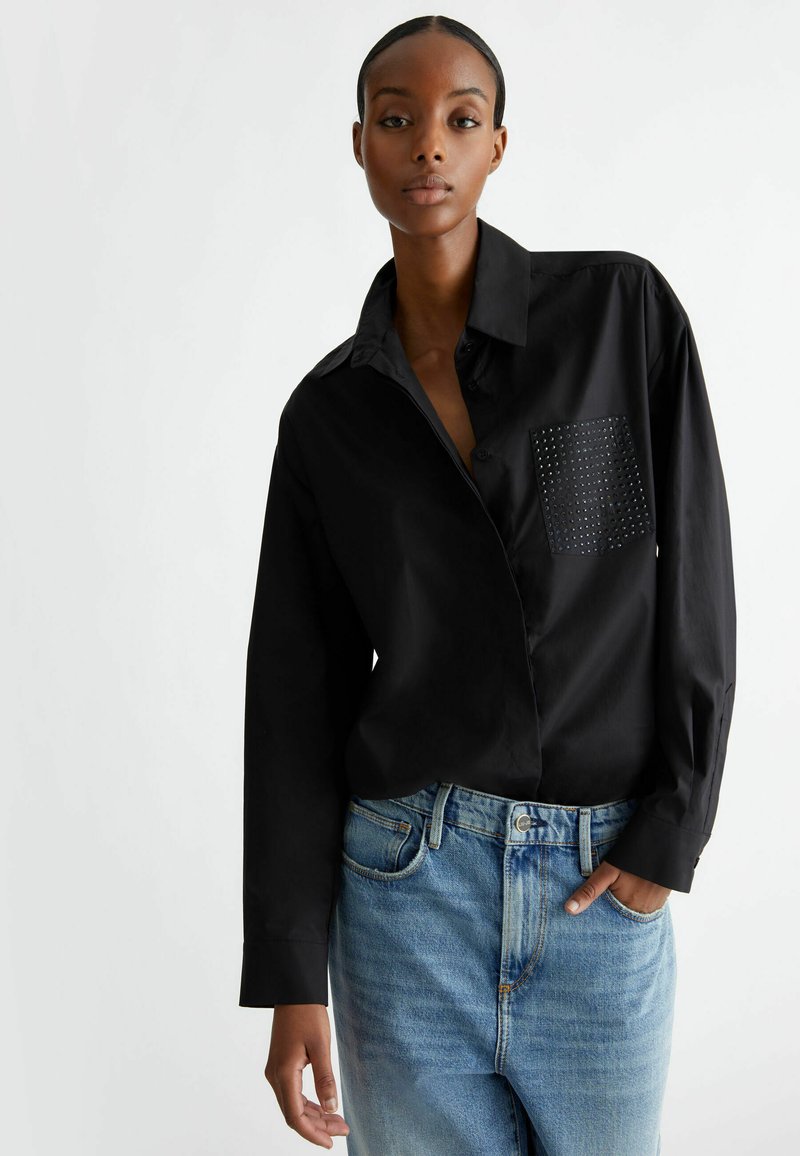 Camisa negra de manga larga con cierre de botones, que presenta un bolsillo texturizado con tachuelas. Se lleva con jeans de tiro alto de color azul claro.
