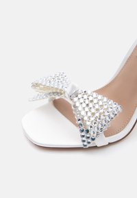 Λευκό σανδάλι με ψηλό τακούνι, ανοιχτή μύτη και μεγάλο φιόγκο διακοσμημένο με διαυγή στρας στην μπροστινή λουρίδα.