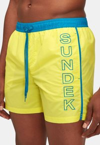 Bañadores amarillos con una cinturilla elástica azul, que presentan un cordón y un atrevido texto "SUNDEK" a lo largo del costado. Material suave y liviano.