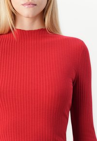 Rød stripete langermet topp med høy hals. Teksturen er myk, og har en tettsittende design som fremhever kroppsformen.