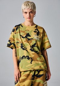 Camiseta de gran tamaño con estampado de camuflaje en verde, marrón y negro, hecha de una tela suave con un cuello redondo y mangas cortas. Acentuada con texto en blanco.