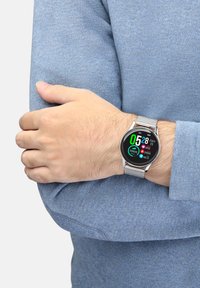 Orologio smart rotondo con display nero, cinturino in rete argento e lettura digitale colorata, indossato su un polso con un maglione blu chiaro.