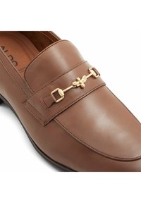 Mocassin en cuir marron avec détail de boucle en métal en forme d'abeille dorée sur la sangle, présentant des coutures le long des bords.