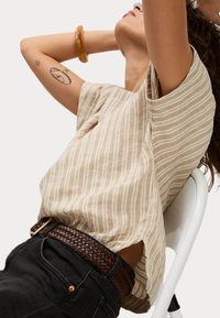 Top de manga corta a rayas beige con un corte holgado. Lleva puestas pantalones de mezclilla oscuros y un cinturón trenzado marrón. La modelo posa frente a un fondo blanco.