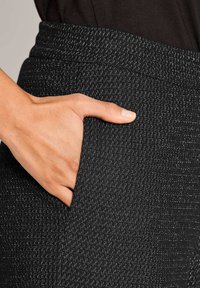 Hand rustend in de voorzak van zwarte, gestructureerde broek met fijne witte stiksels.