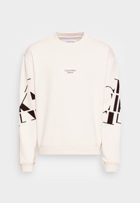 Sweatshirt Calvin Klein Jeans off-white avec col rond, petit logo sur la poitrine et grand texte graphique noir sur les deux manches.