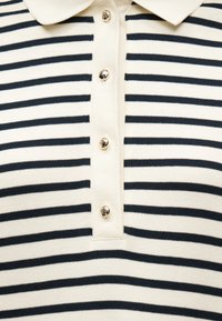 Πόλο πουκάμισο με ρίγες σε κρεμ και μαύρο χρώμα, με γιακά και τέσσερα χρυσά κουμπιά κατά μήκος της εμπρόσθιας λωρίδας.