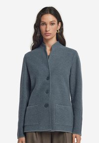 Grå texturerad cardigan med ståkrage, fyra framsnappar och två fickor. Den har en boxig passform med lång ärm.