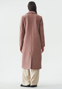Cappotto lungo rosa realizzato in un tessuto strutturato, caratterizzato da una fessura posteriore, colletto a punta e vestibilità sartoriale, abbinato a pantaloni larghi di colore chiaro.
