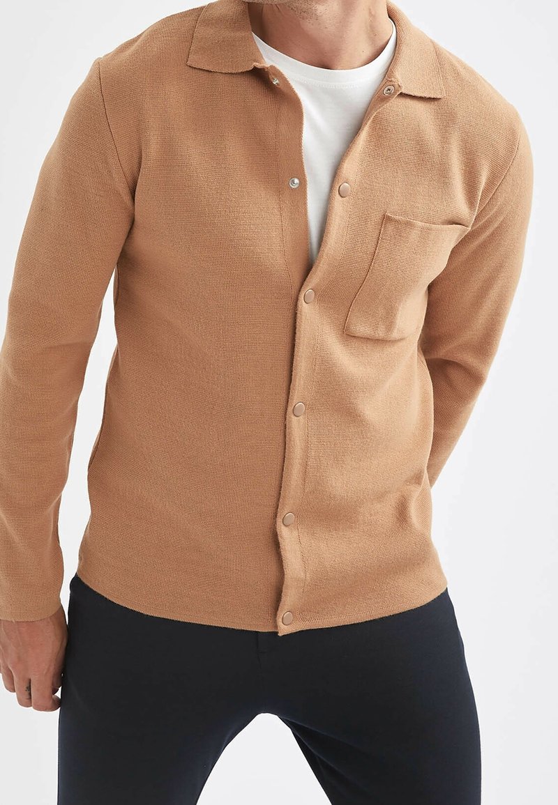Homme portant un cardigan brun clair boutonné sur une chemise blanche et un pantalon noir sur un fond clair uni.