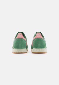 Green suede sneakers with pink accents, featuring a textured sole and a prominent heel tab. Back view shows two shoes side by side.