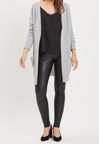 Světle šedý pletený kardigan s otevřeným předním dílem, dlouhými rukávy a kapsami, přetáhnutý přes černý top s krajkovým lemem, v kombinaci s černými koženými legínami a podpatky.