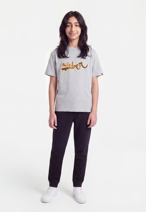 Jong persoon met lang donker haar, een grijs T-shirt met print, zwarte broek en witte sneakers, staand tegen een witte achtergrond.