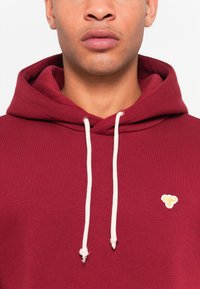Burgundy hoodie with white drawstrings, featuring a small yellow logo on the left chest. Material appears soft with a smooth texture.