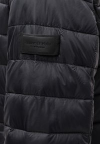 Juodas pūkinis švarkas su siuvinėta tekstūra ir vidiniu etiketu. Sušildymo dalys sujungtos siūlėmis ir minimalistinis, elegantiškas dizainas.